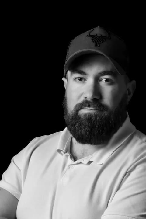 Kevin Machy, portrait en noir et blanc sur fond noir, bras croisés, casquette au logo de bois de cerf