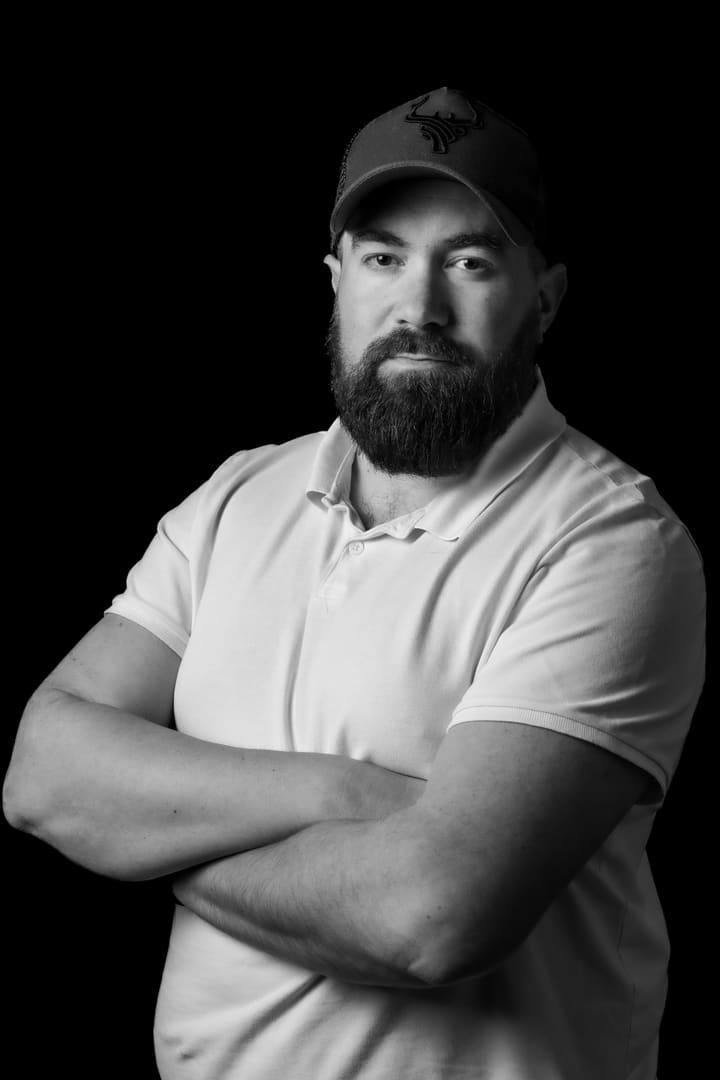 Kevin Machy, portrait en noir et blanc sur fond noir, bras croisés, casquette au logo de bois de cerf