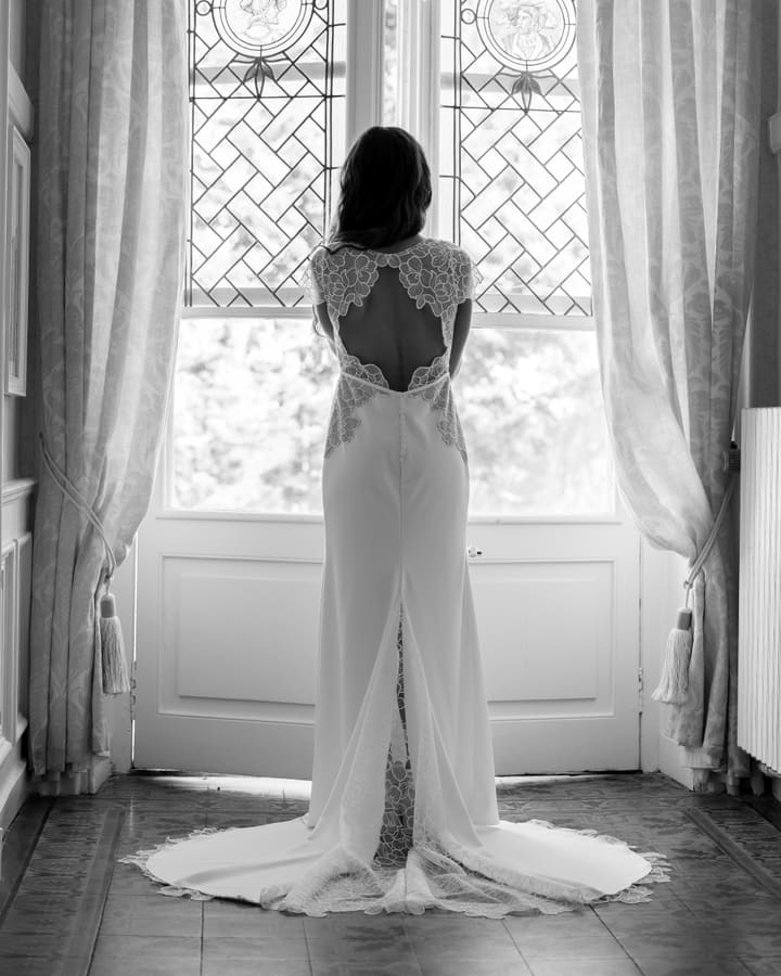 La mariée de dos devant une fenêtre, le dos de sa robe ouvert sur de la dentelle.