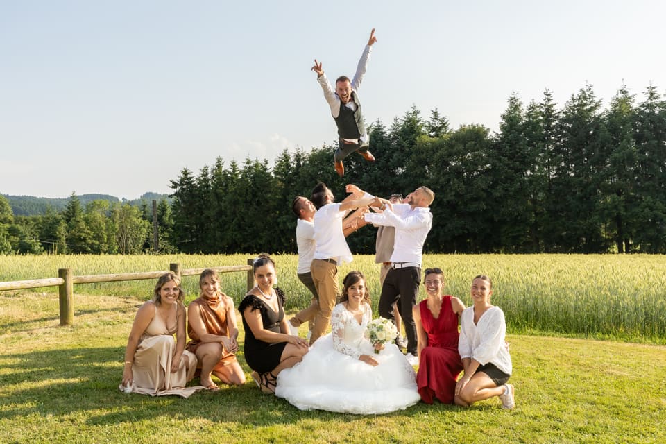 Les mariés et leurs invités assis dans un pré, l’un d’eux saute bras levés.