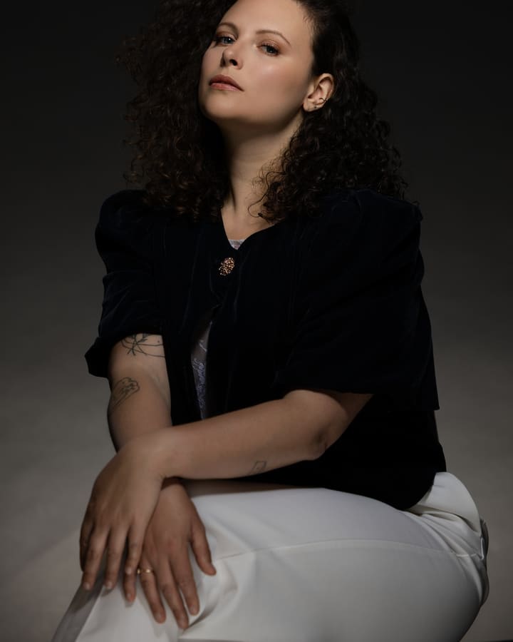 Une femme assise de trois quarts, veste noire et pantalon blanc, sur fond gris.